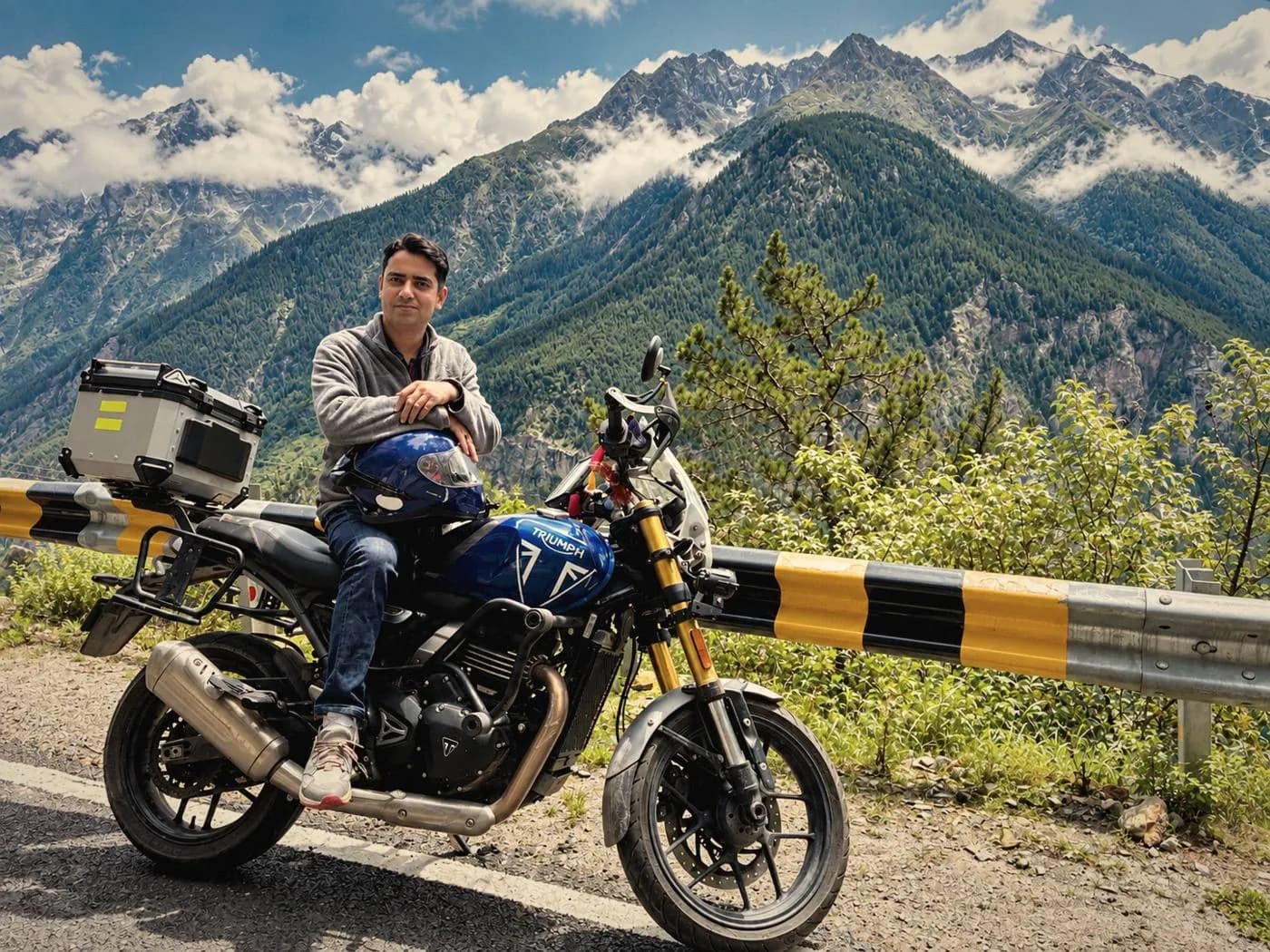 One of HimVeda’s four founders seated on a blue Triumph motorcycle beside a mountain road, with forested Himalayan peaks and clouds behind him
